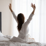 Happy young woman stretching waking up sitting in cozy bed